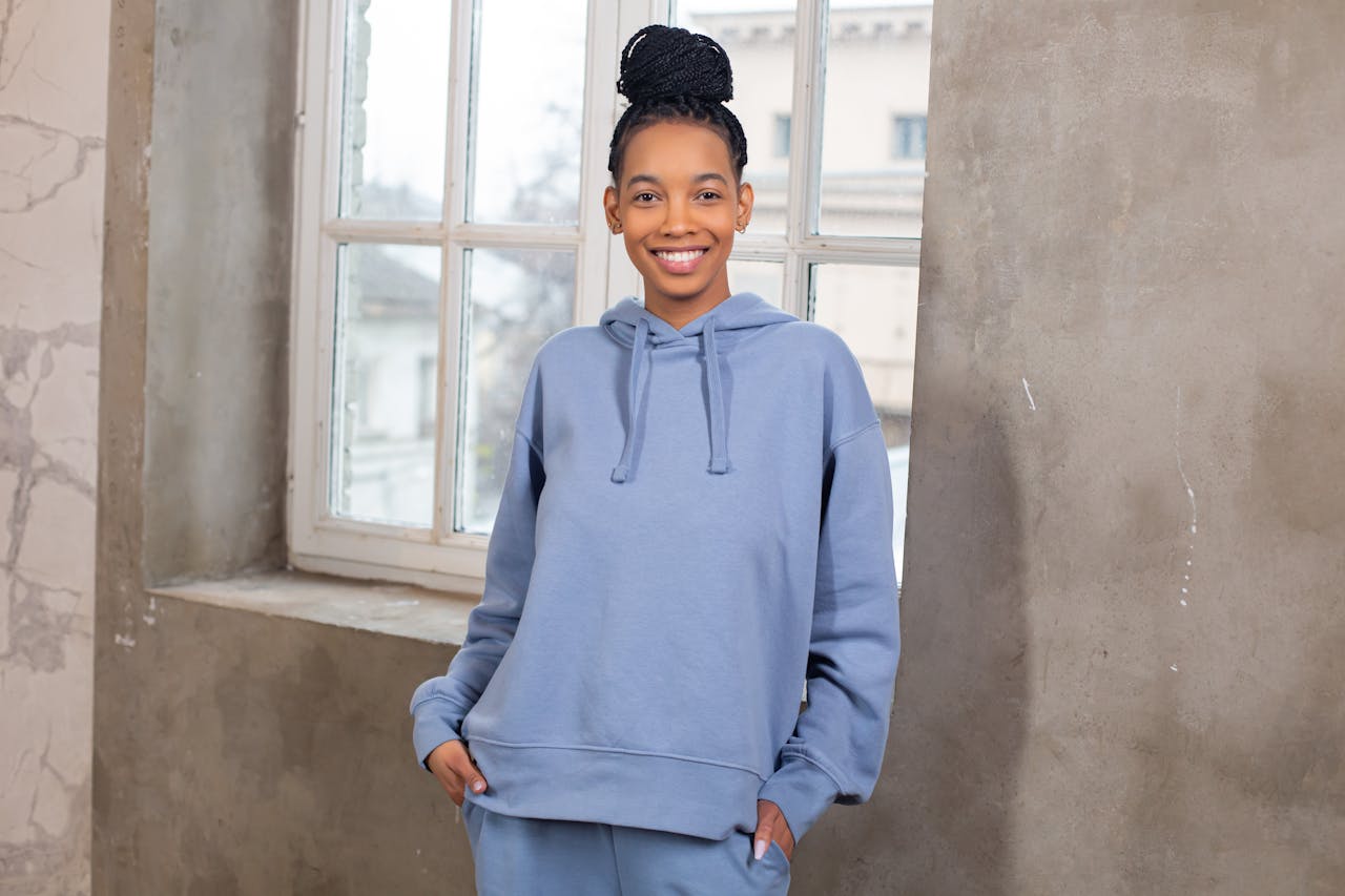 home-hero Cheerful attractive African American female wearing blue cozy hoodie standing with hands in pockets and looking at camera while standing near window in light room